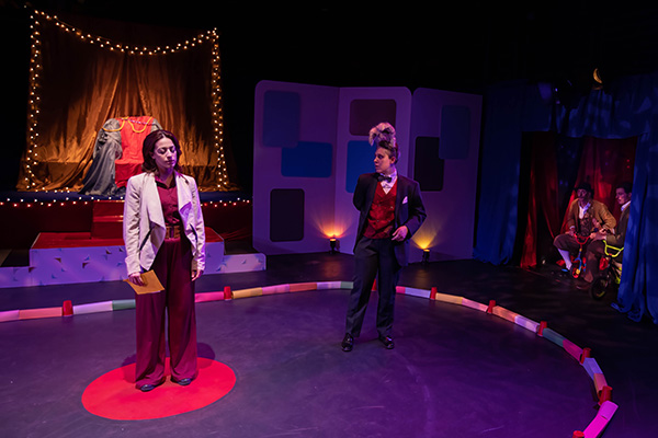 Photo of Kat Gonzalez as Sheela and Dani Martineck as The Hare standing in a circus ring in front of a throne surrounded by Christmas lights. They are preparing to ask a question of four male contestants sitting in a curtained alcove to their left on children's bikes.