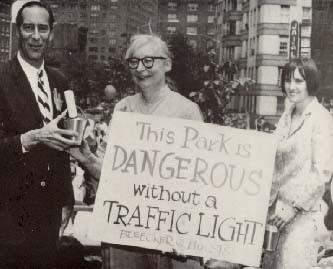 Jane Jacobs being interviewed at a protest; she has a sign that reads This Park is Dangerous Without a Traffic Light!